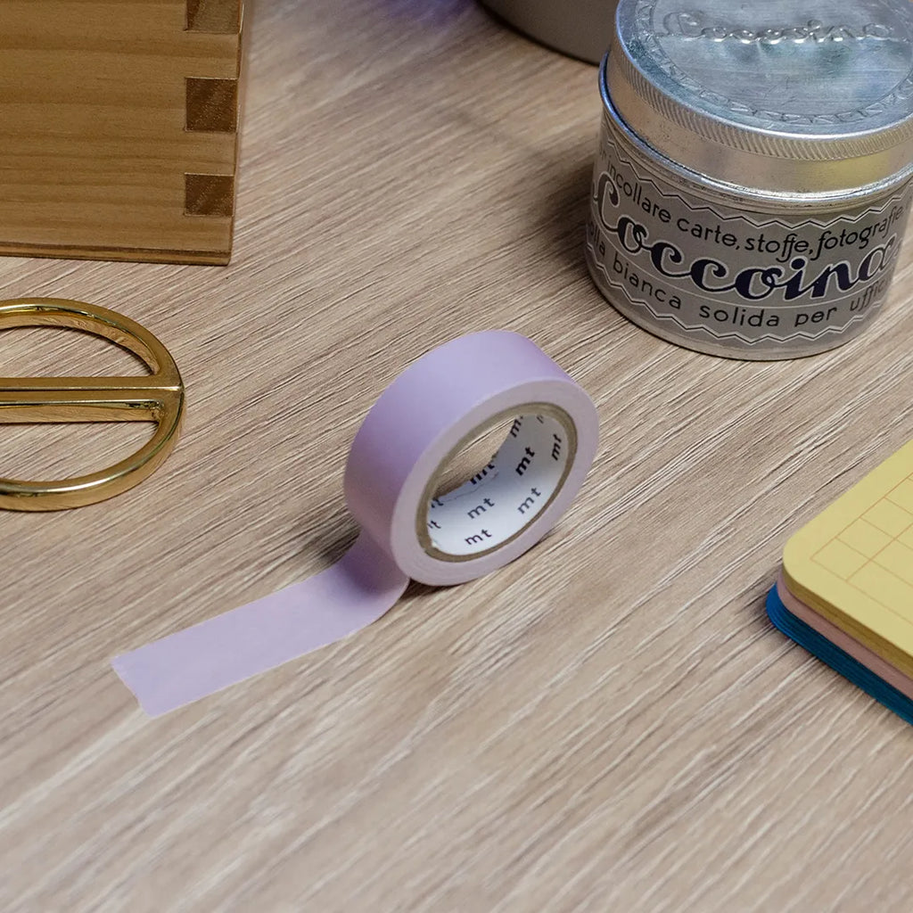 Rouleau de masking tape violet pastel posé sur un bureau en bois. Avec des objets autour : colle, foglietti, boîte en bois, ciseaux. 