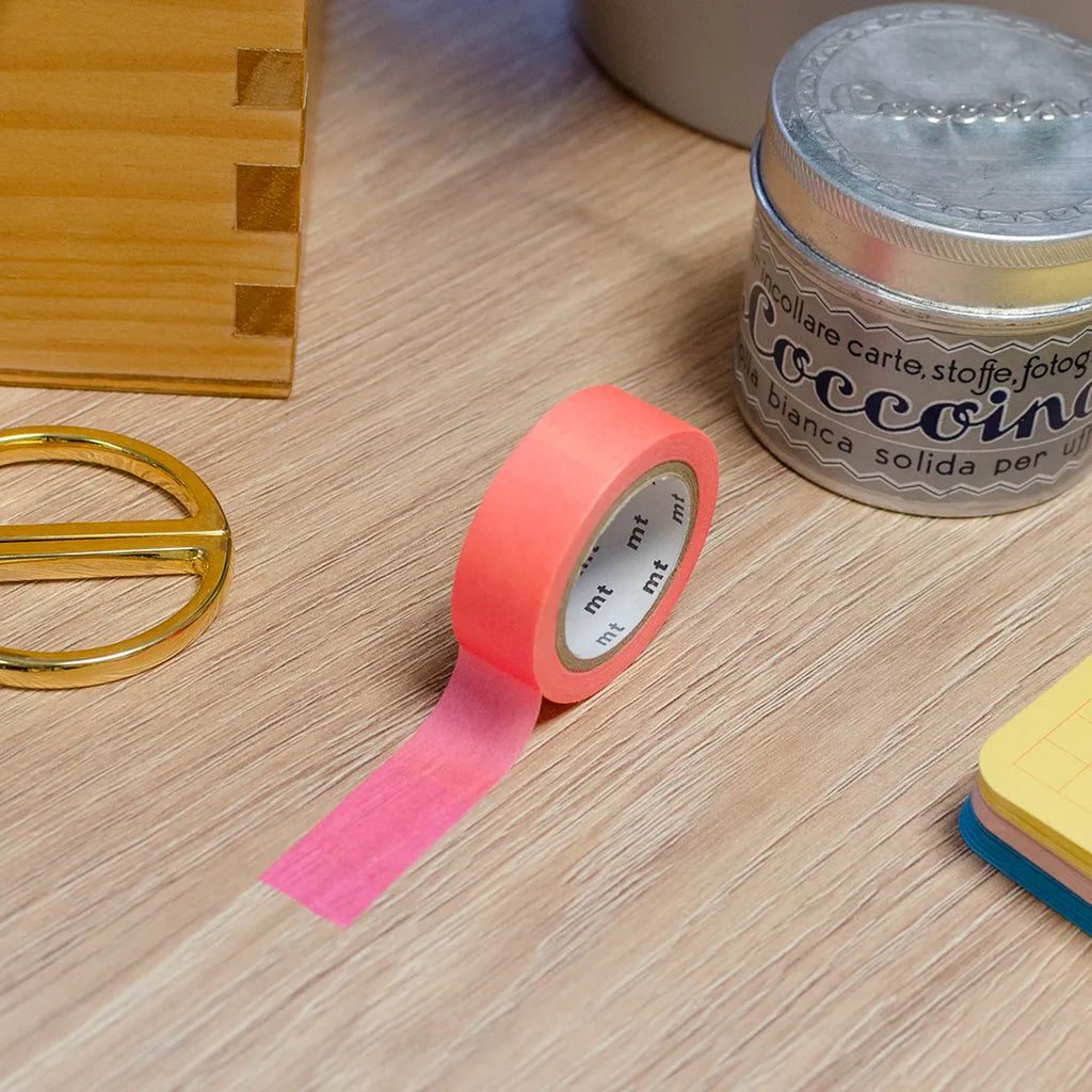 Rouleau de masking tape dégradé rose-orange posé sur un bureau en bois. Avec des objets autour : colle, foglietti, boîte en bois, ciseaux. 