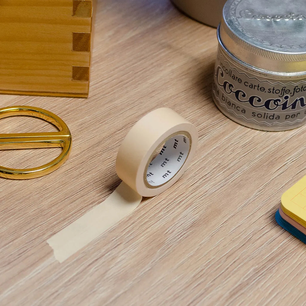 Rouleau de masking tape couleur beige, posé sur un bureau en bois. Avec des objets autour : colle, foglietti, boîte en bois, ciseaux. 