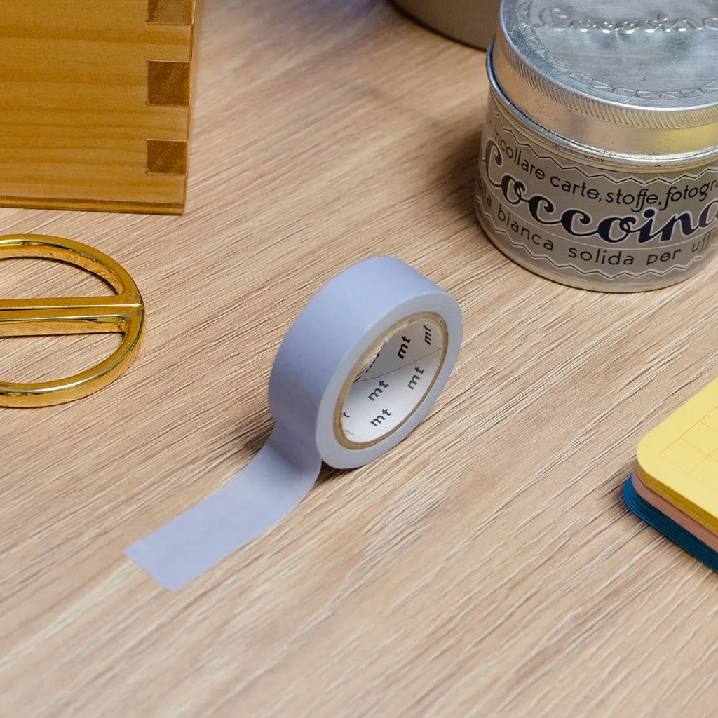 Rouleau de masking tape bleu-violet pastel posé sur un bureau en bois. Avec des objets autour : colle, foglietti, boîte en bois, ciseaux. 