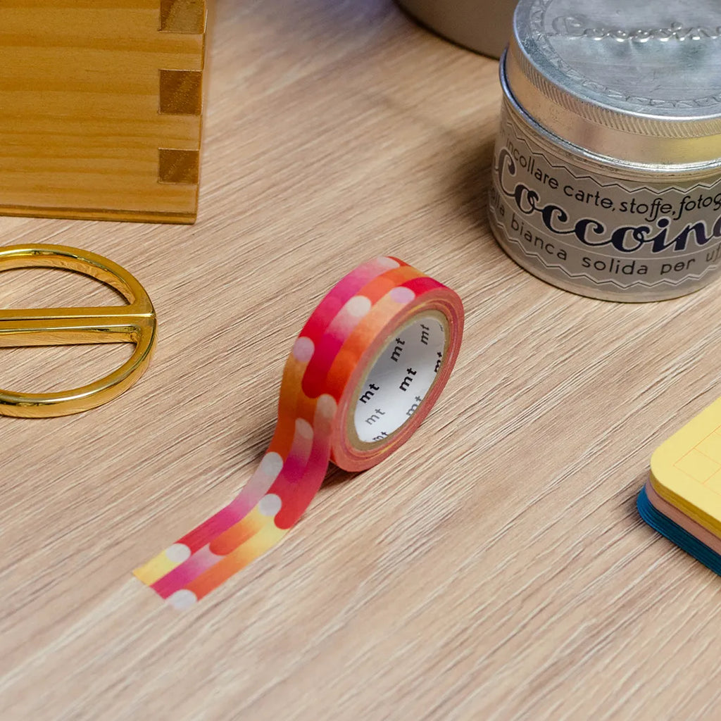 Rouleau de masking tape à motif rose, orange, jaune, blanc, rouge, violet, posé sur un bureau en bois. Avec des objets autour : colle, foglietti, boîte en bois, ciseaux. 