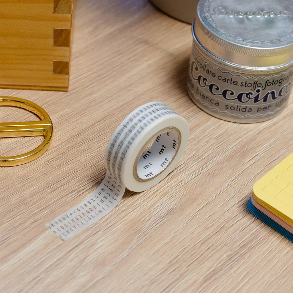 Rouleau de masking tape avec des numéros et des chiffres, posé sur un bureau en bois. Avec des objets autour : colle, foglietti, boîte en bois, ciseaux. 