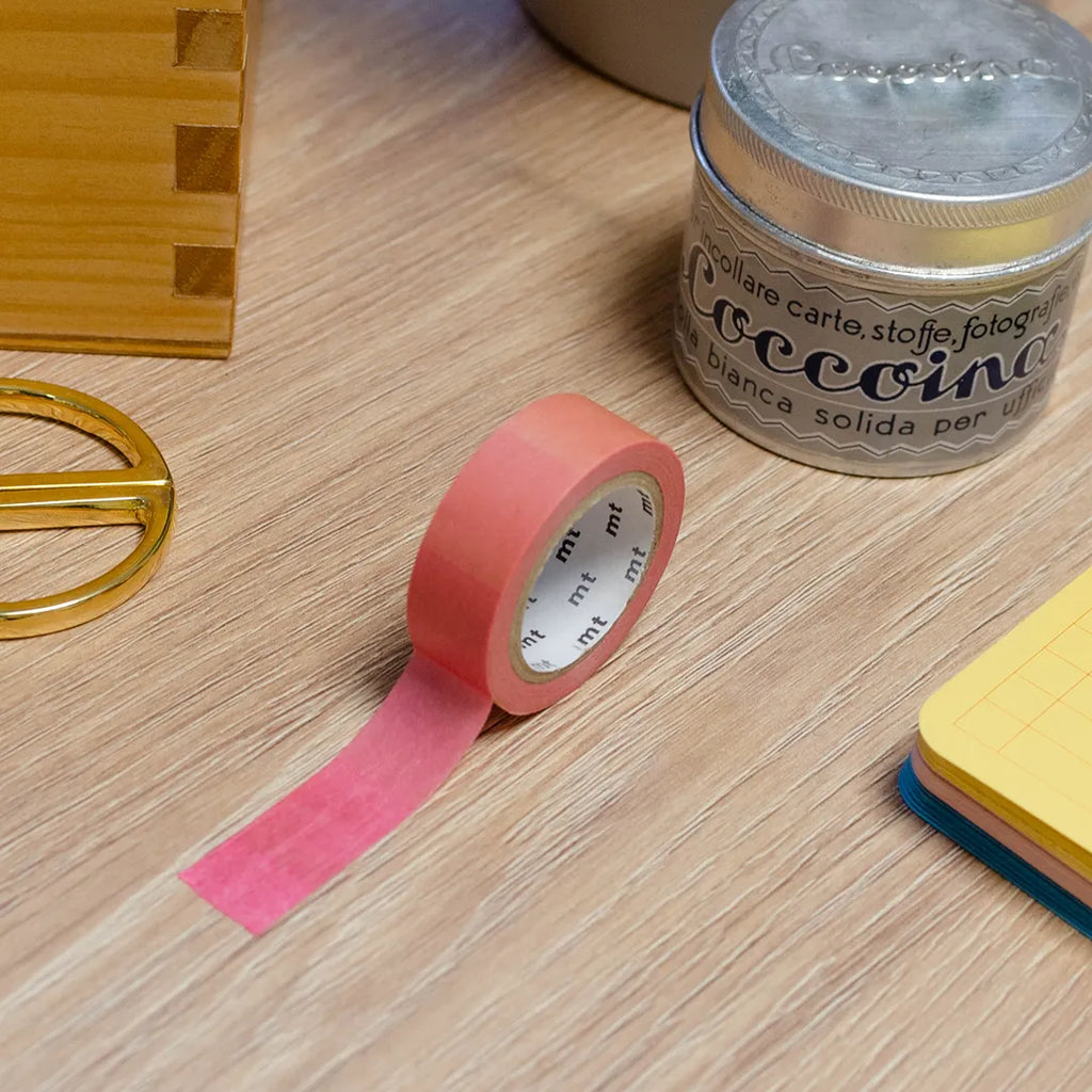 Rouleau de masking tape dégradé violet, rose, orange, jaune, posé sur un bureau en bois. Avec des objets autour : colle, foglietti, boîte en bois, ciseaux. 