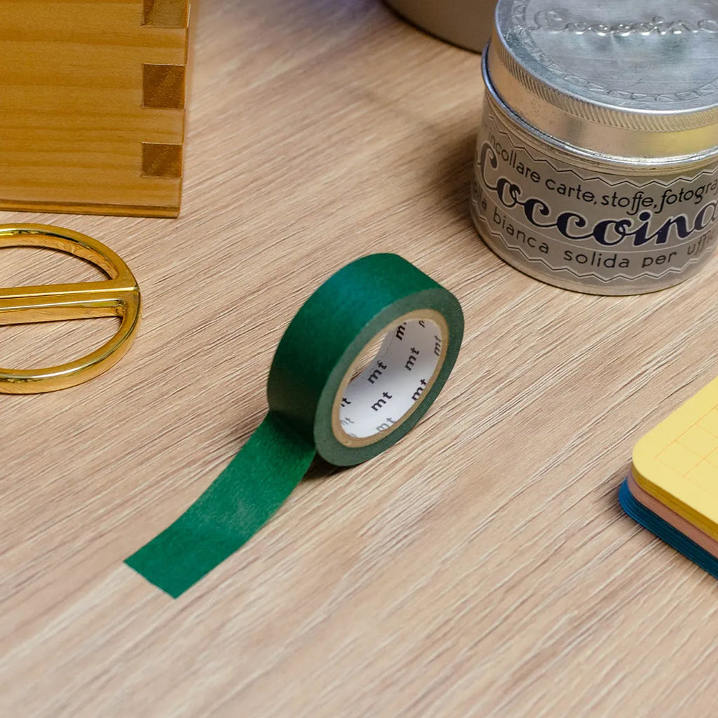 Rouleau de masking tape vert sapin posé sur un bureau en bois. Avec des objets autour : colle, foglietti, boîte en bois, ciseaux. 