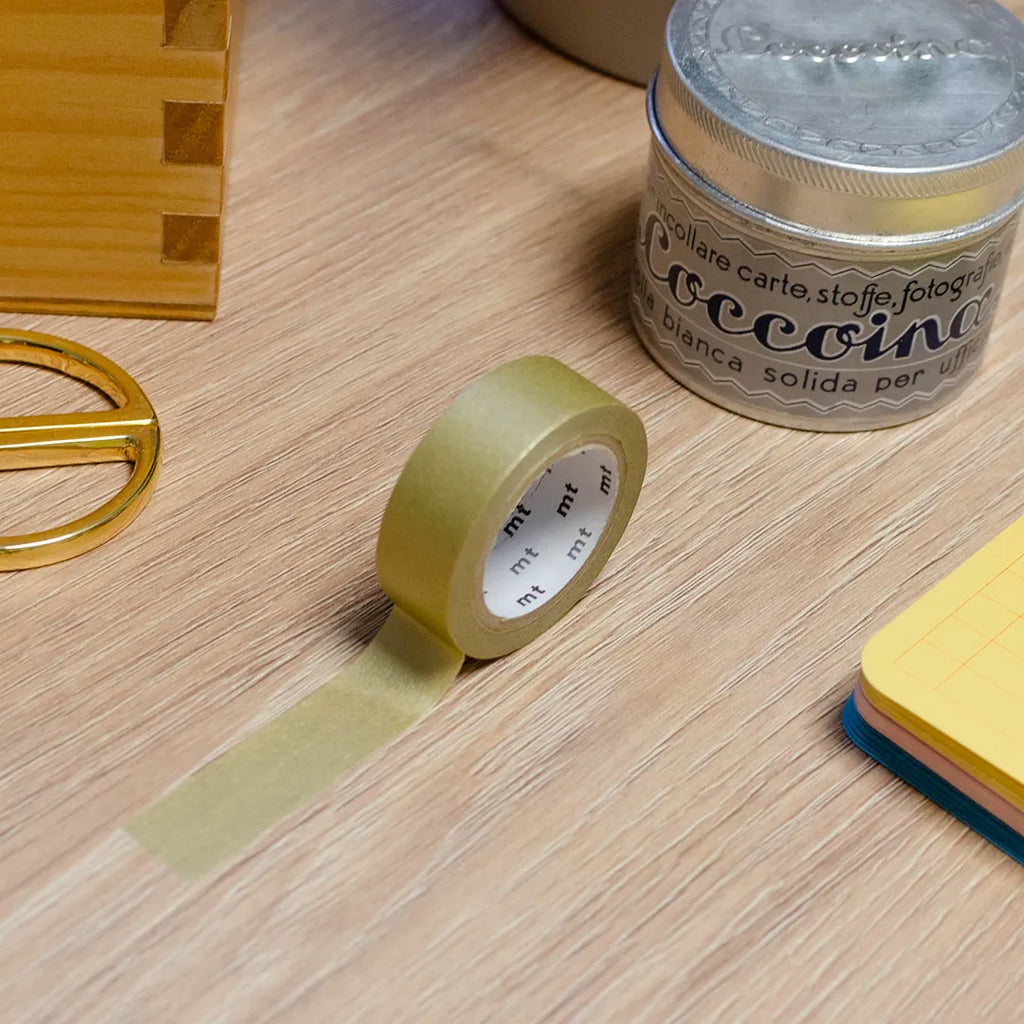 Rouleau de masking tape couleur or, posé sur un bureau en bois. Avec des objets autour : colle, foglietti, boîte en bois, ciseaux. 