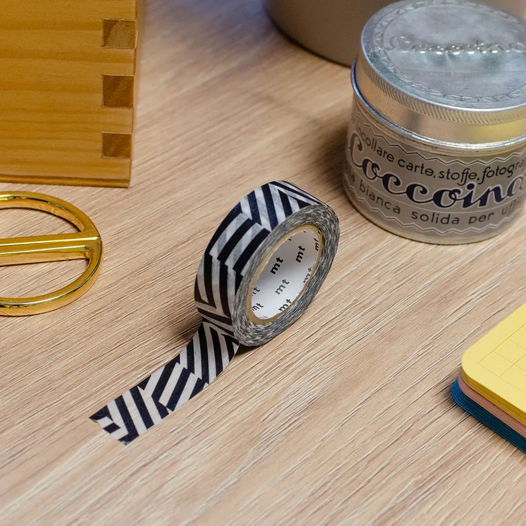 Rouleau de masking tape à motif en ligne noir et blanc, posé sur un bureau en bois. Avec des objets autour : colle, foglietti, boîte en bois, ciseaux. 